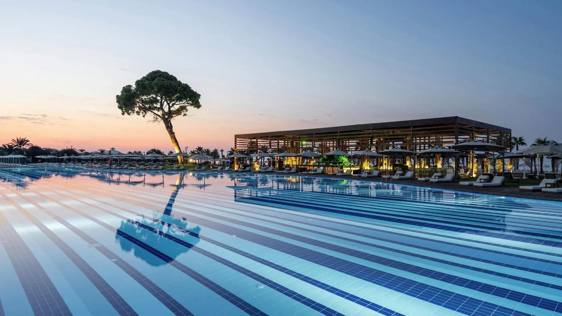 Rixos Premium Belek - The Land Of Legends Access Hotel Exterior photo This is a beautiful, serene photograph capturing a luxury resort area around twilight or early morning.

The foreground is dominated by a large, modern swimming pool. The water surface is highly reflective, and the pool itself features a striking des